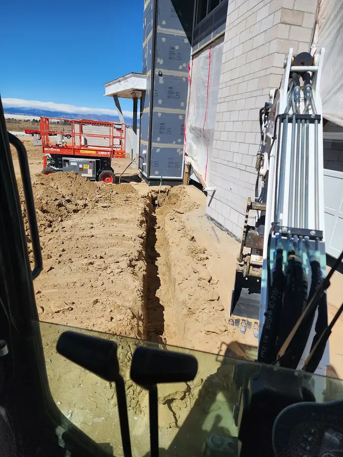 View from the excavator cab of a narrow utility trench being dug beside a new home