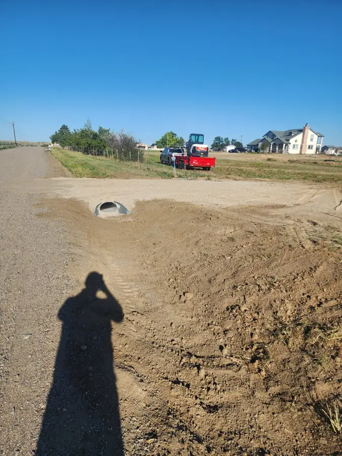 A new culvert under a gravel driveway entrance off a rural road, with a red excavator in the distance