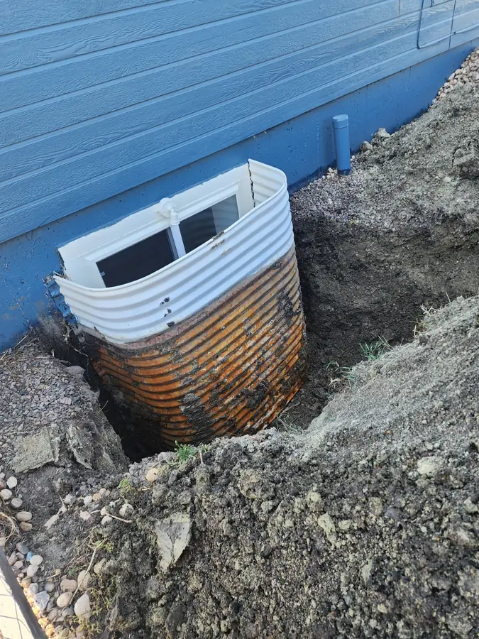 An excavated egress window well beside a blue house foundation, with gravel at the bottom