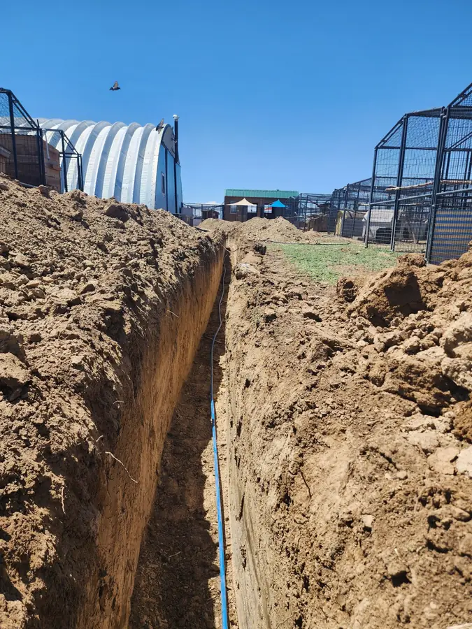 A deep, straight utility trench with a blue line at the bottom beside metal outbuildings